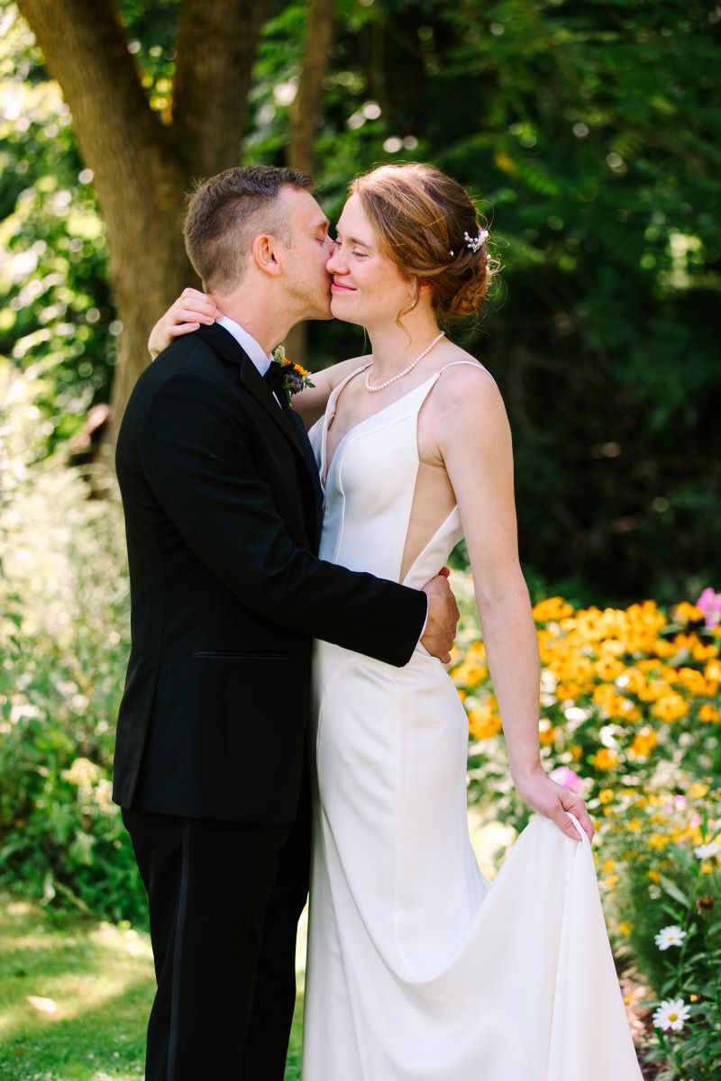 A woman in a white wedding dress with one hand is holding up the side of her white dress while her husband who is wearing a black tuxedo leans in to kiss her 