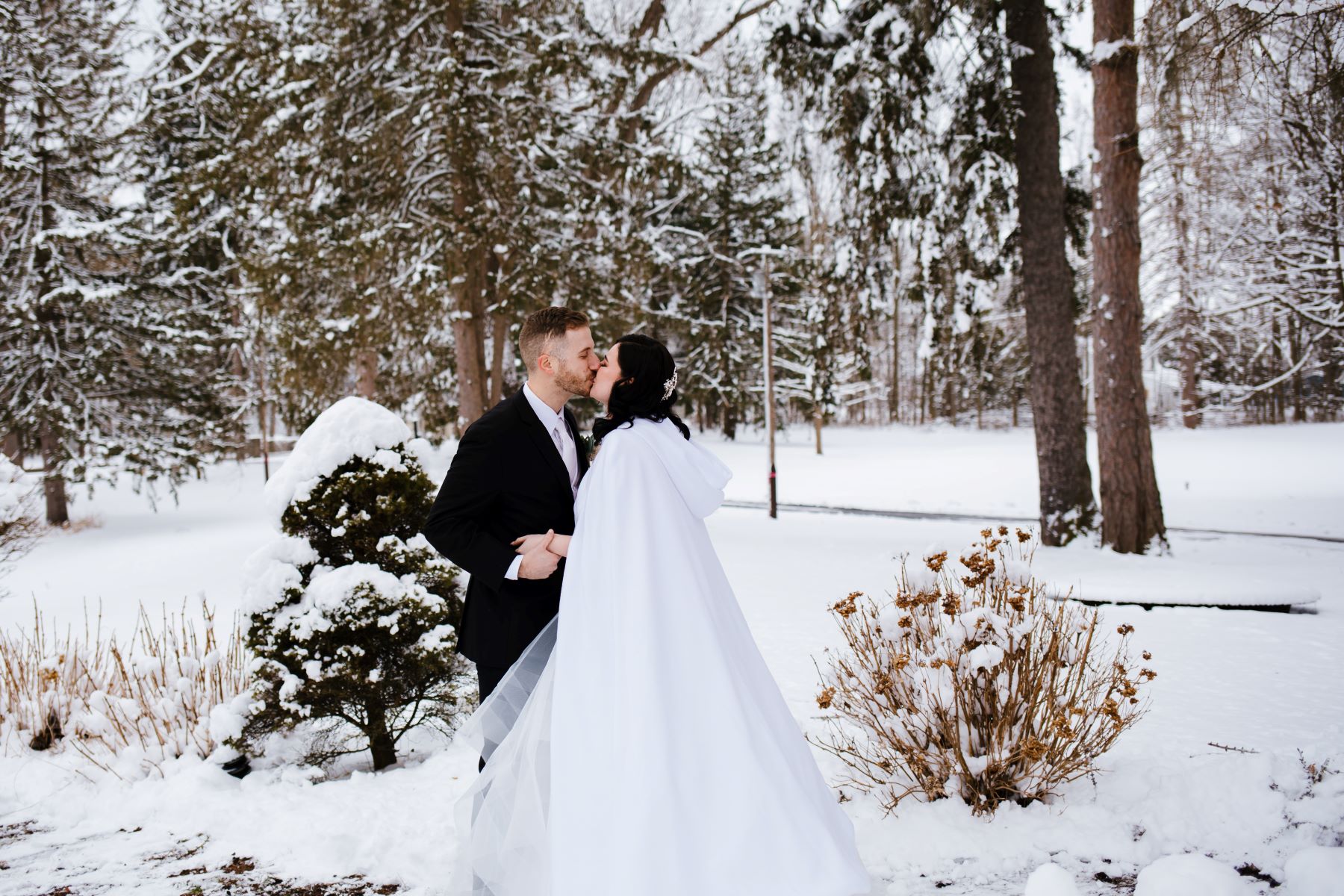 A couple kissing in the snow the woman is wearing a white wedding cape and the man is wearing a black tuxedo and behind them are pine trees covered in snow and snow on the ground