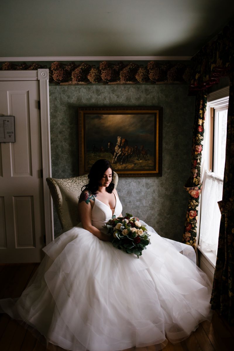 A woman in a white wedding dress sitting in a chair and holding a bouquet of flowers next to a window 