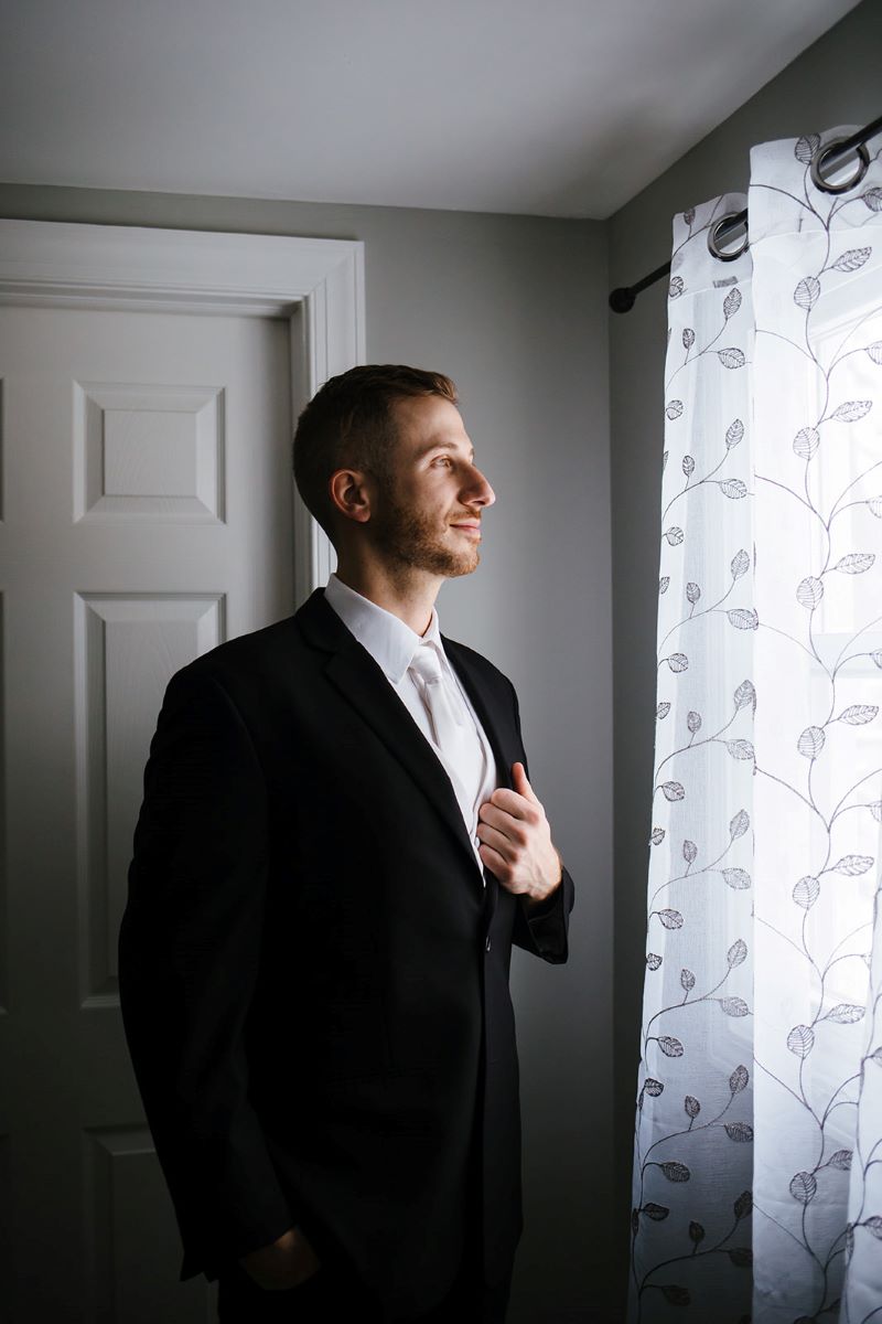 A man who is wearing a black tuxedo looking out a window 