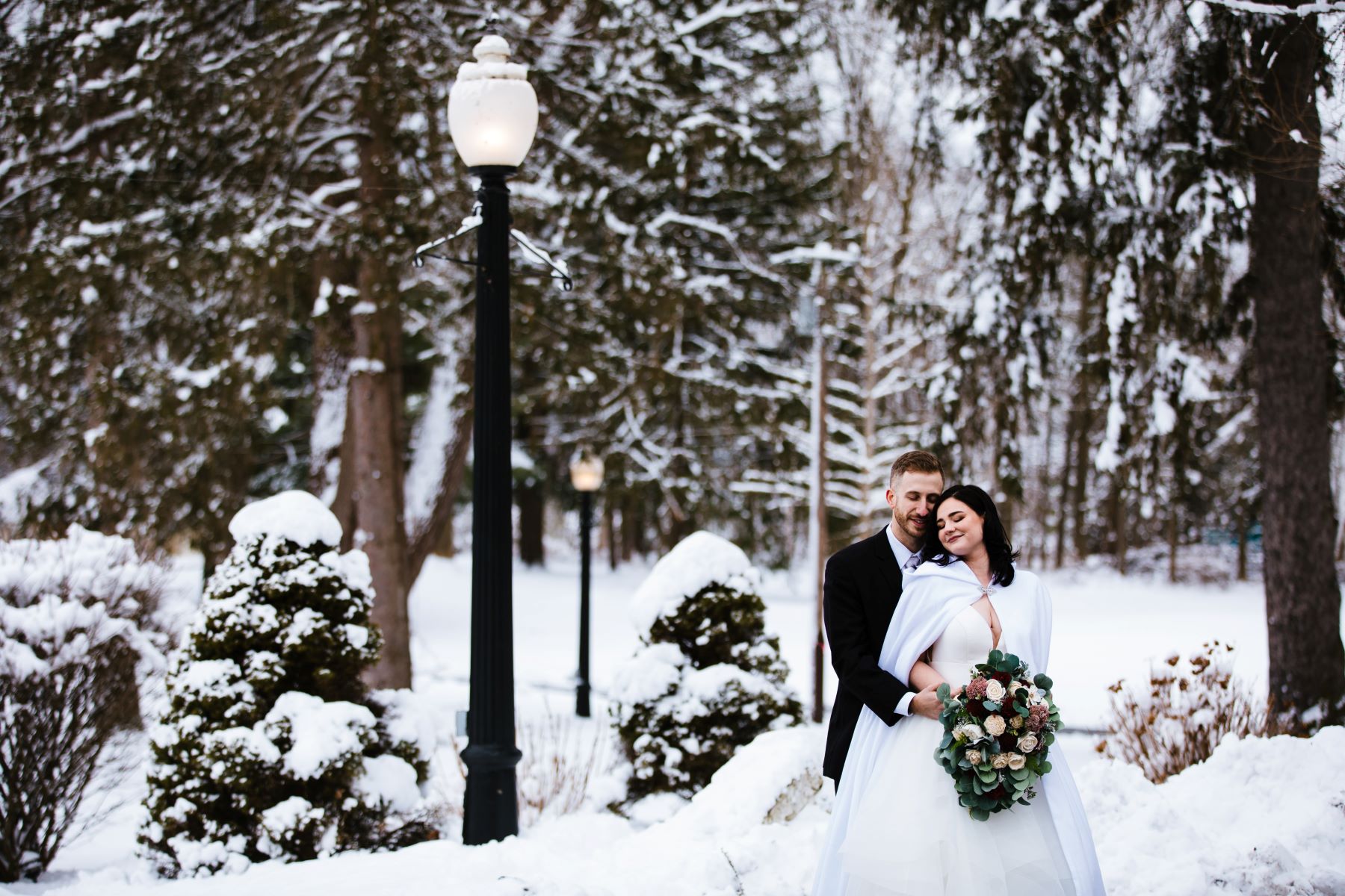 A woman in a white wedding dress is holding a bouquet of flowers and her partner who is wearing a black tuxedo is hugging her from behind and there is snow on the ground behind them and large green pine trees and lamposts