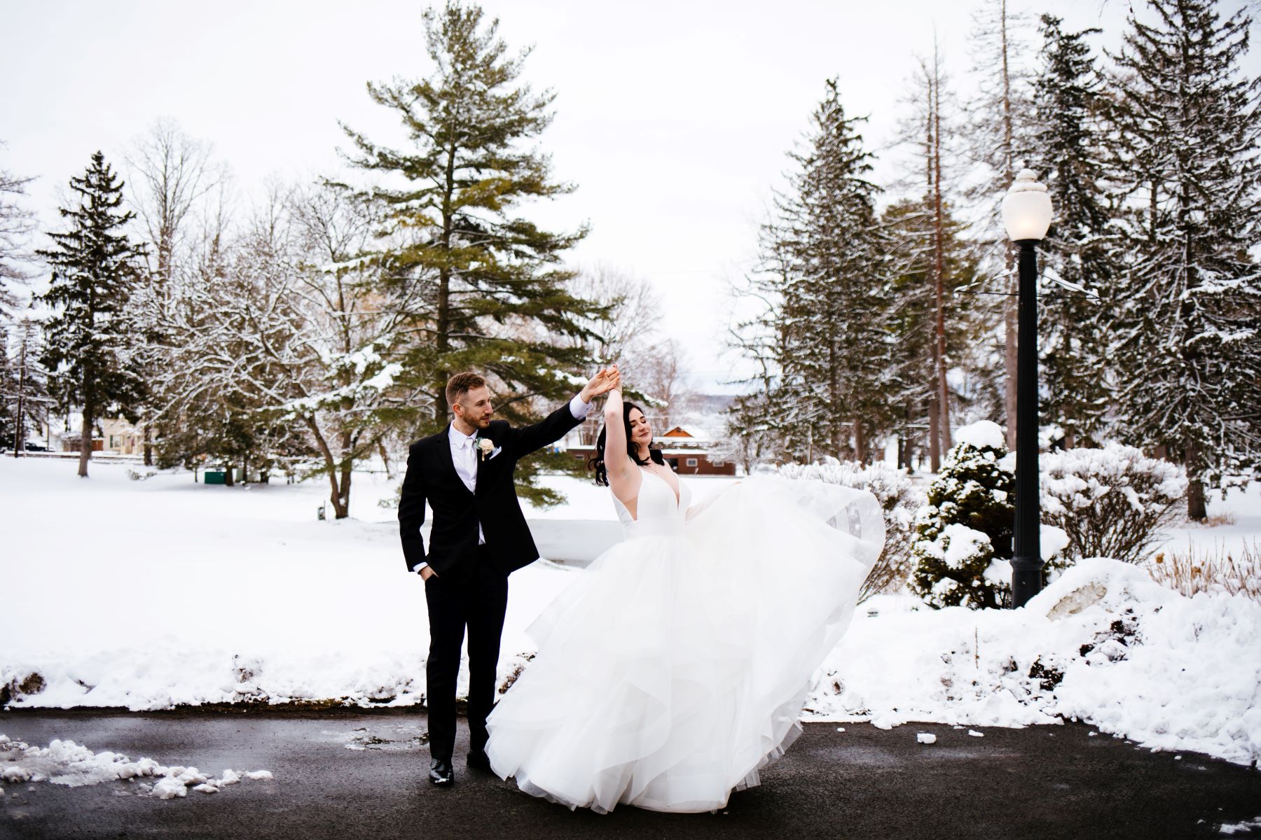 A woman in a white dress is being spun around by her partner who is wearing a black tuxedo and a  boutonniere and a white button up and tie the woman is the wedding dress is lifting up one side of her wedding dress in the air and there is snow on the ground behind them 