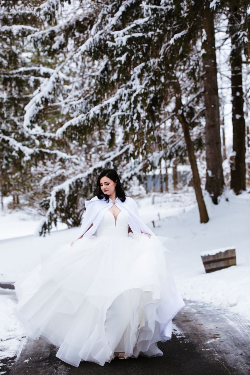 A woman is walking and holding up her white wedding dress in the snow she is also wearing a white wedding cape 