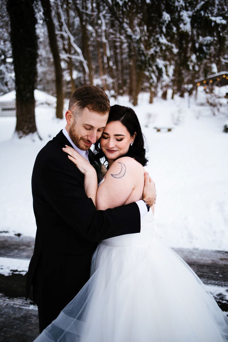 A woman in a white wedding dress with a moon tattoo is hugging her partner who is wearing a black tuxedo 