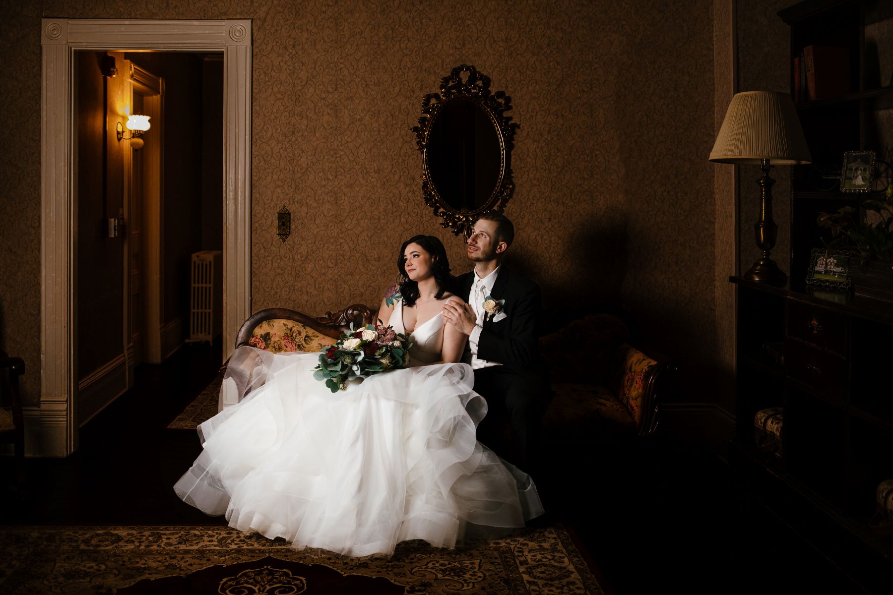 A woman in a white wedding dress is sitting next to her partner who is wearing a black tuxedo on a couh and she is holding a bouquet of flowers and they are both looking off to the side