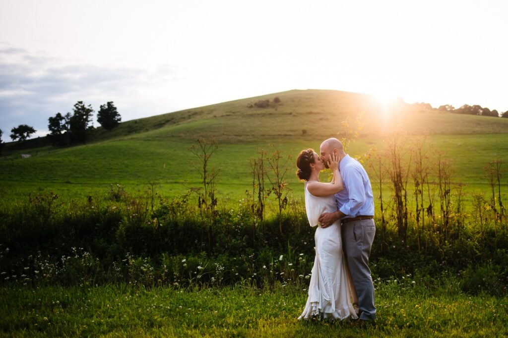 The bride and groom share a kiss in a green field as the sun sets behind a hill.