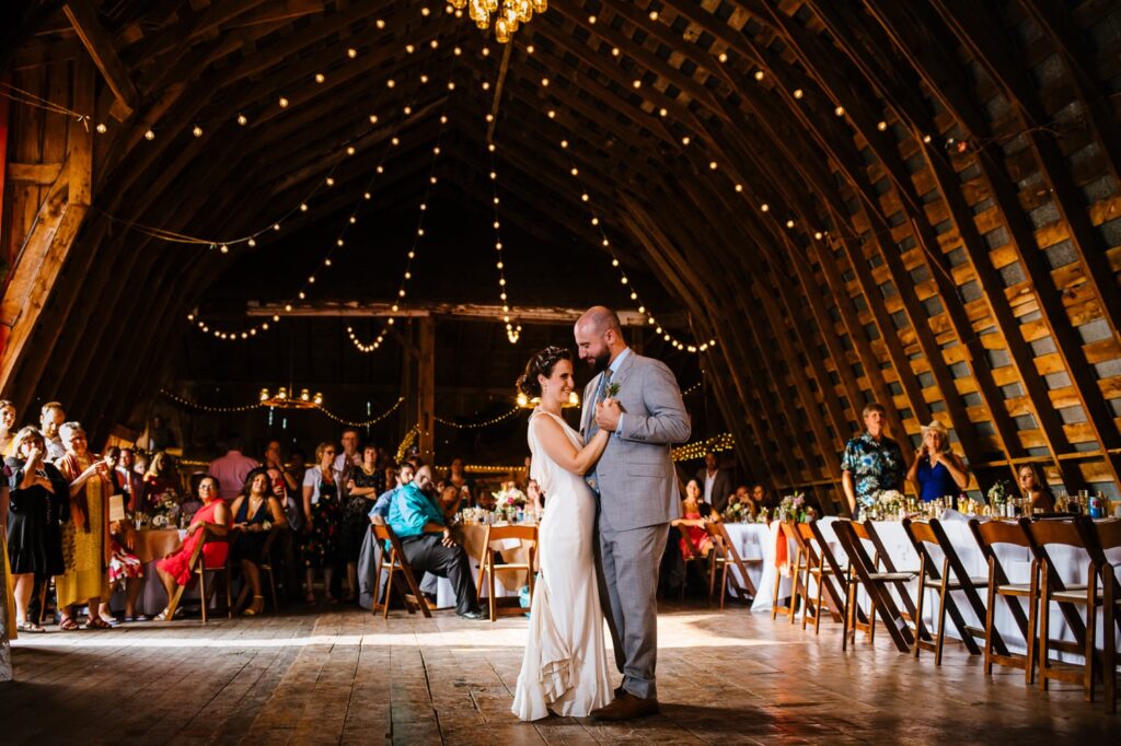 The couple shares their first dance under warm string lights and a wooden vaulted ceiling, surrounded by wedding guests.