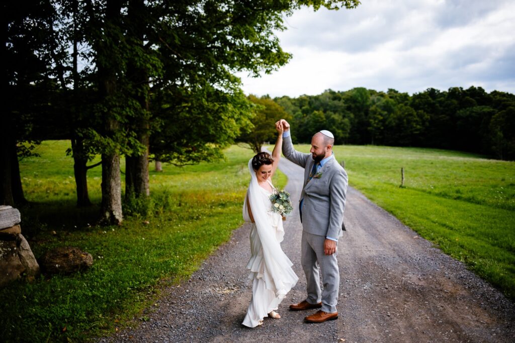 The groom twirls the bride on a gravel lane surrounded by green fields and tall trees. 