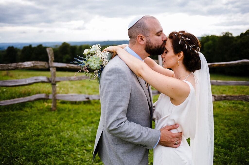 The bride and groom embrace next to a wooden fence with hills in the background.