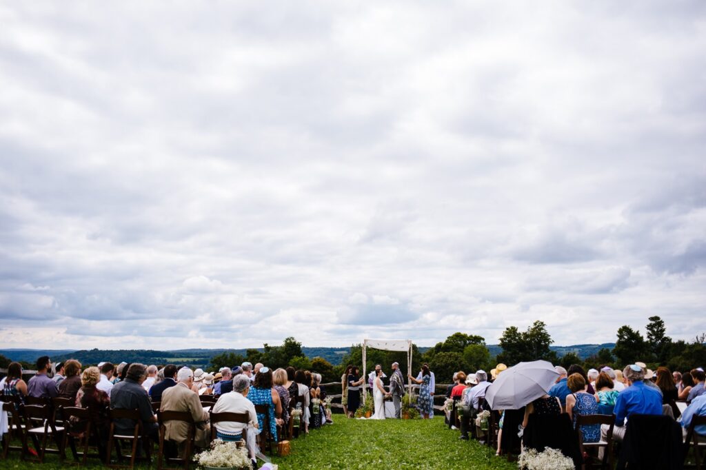 An outdoor wedding ceremony with the couple surrounded by guests seated on either side. The sky is overcast.