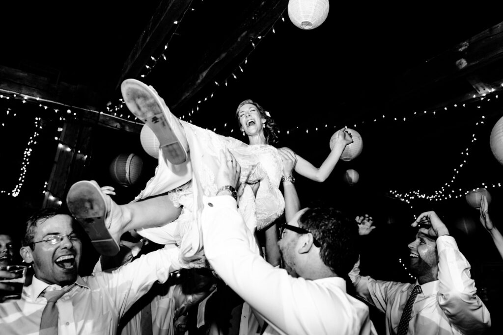 The bride being lifted high by laughing guests, her gown flowing while she smiles.