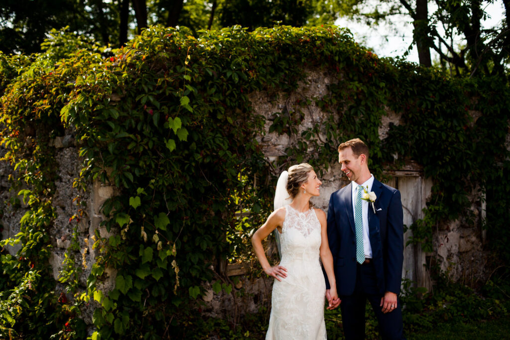 The bride and groom stand hand-in-hand in front of an ivy-covered stone wall.