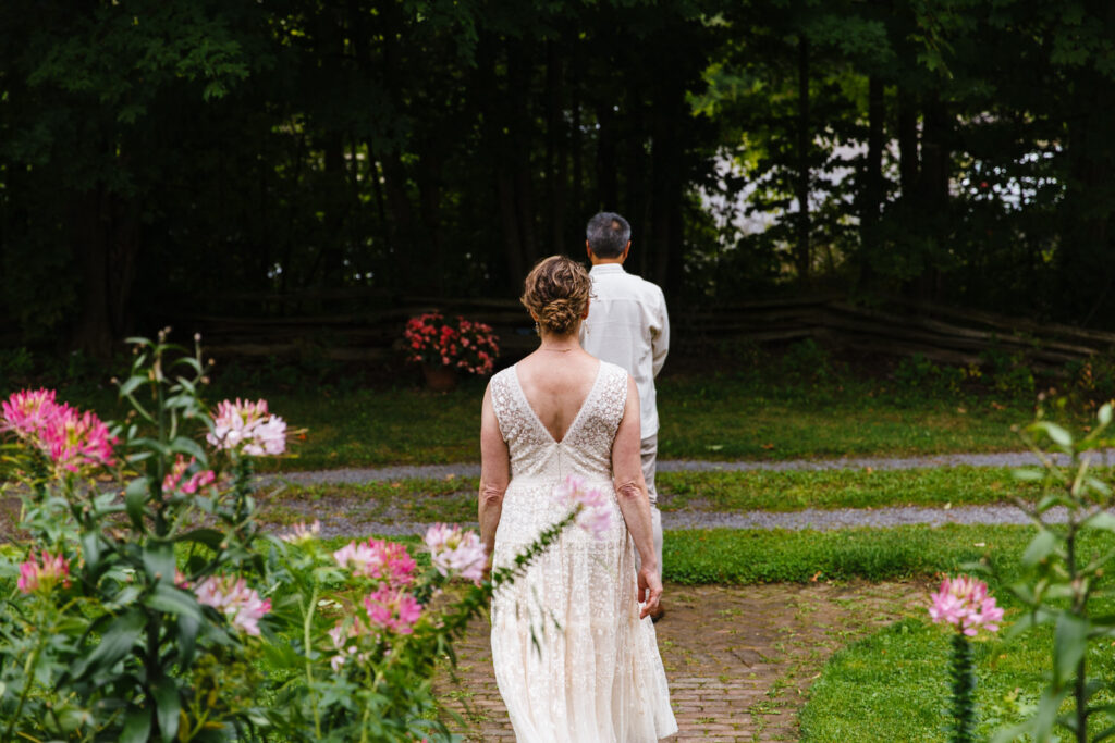 A bride in a lace gown walks along a brick path toward her groom, who stands with his back turned in a garden.