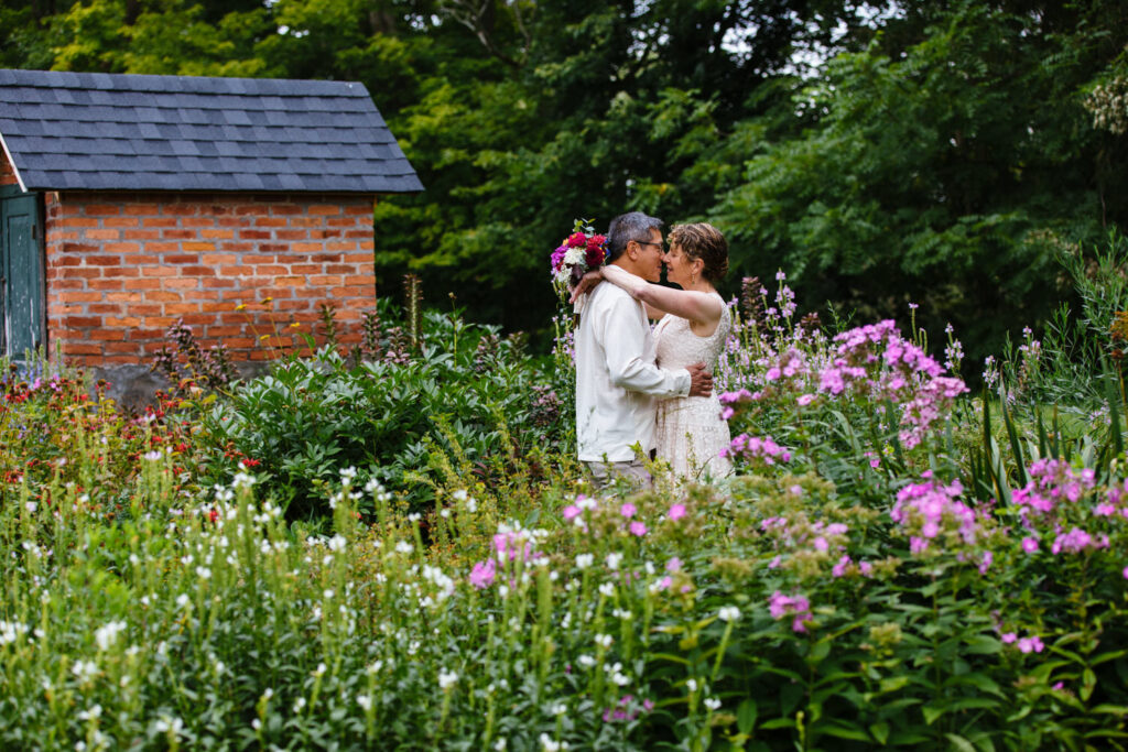 The bride and groom embrace in a colorful garden filled with pink, purple, and white flowers, with a brick building in the background.