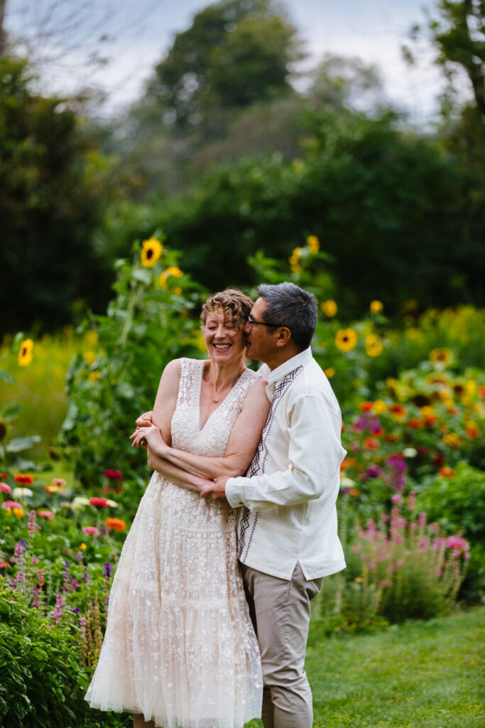 The bride and groom laugh and hug in a field surrounded by bright sunflowers and wildflowers.