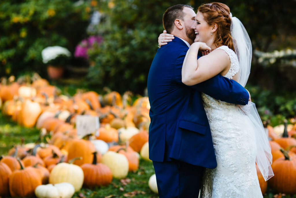 The couple embraces in front of a display of orange and white pumpkins, with the bride in a lace gown and the groom in a navy blue suit. 