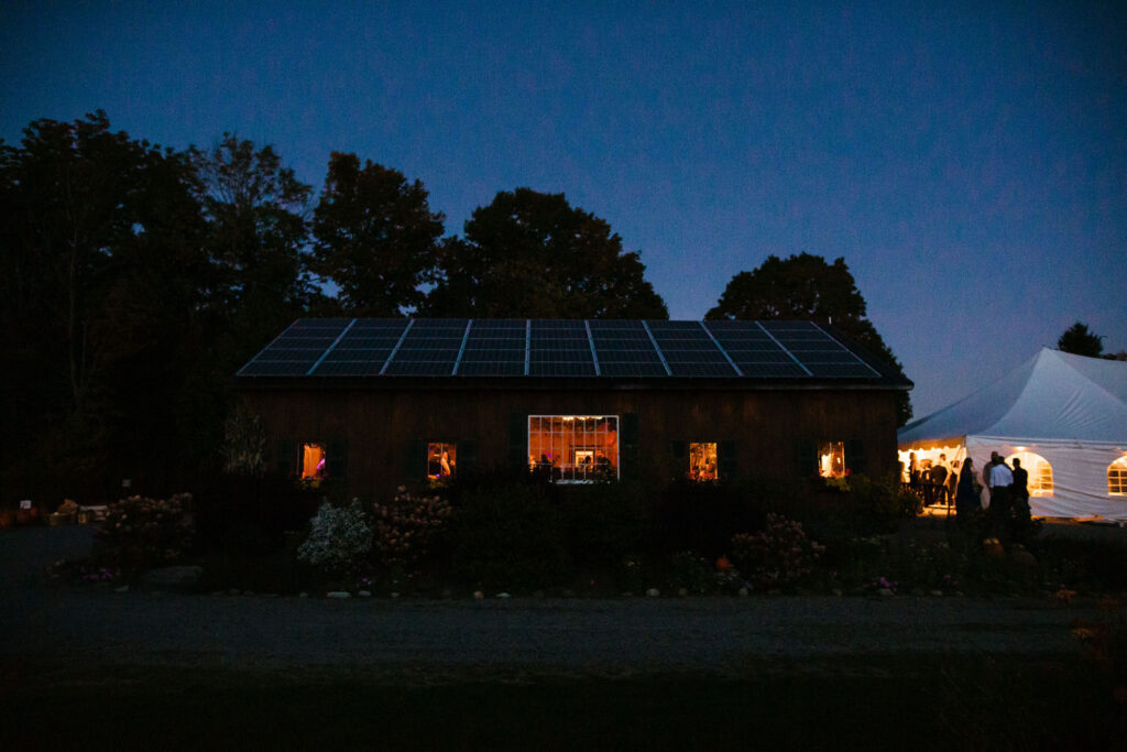 A rustic barn adorned with solar panels is softly lit from the inside, with a glowing white tent nearby hosting wedding festivities. 