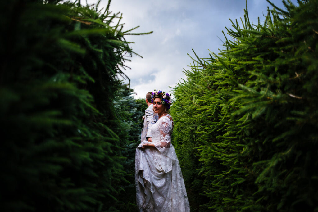 The bride, wearing a lace dress and floral crown, leads the groom through a maze, with the groom in a light suit behind her.