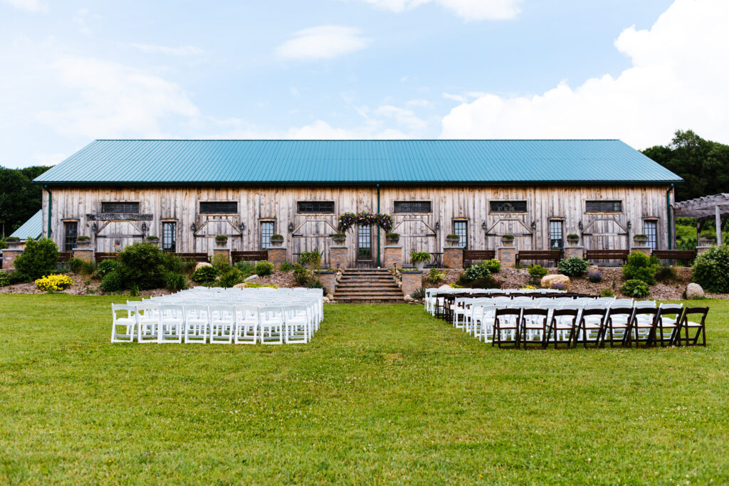 Rows of white chairs are arranged on a green lawn in front of a large wooden barn with a teal roof. The barn features stone steps, flowers, and a rustic floral arch in the center.