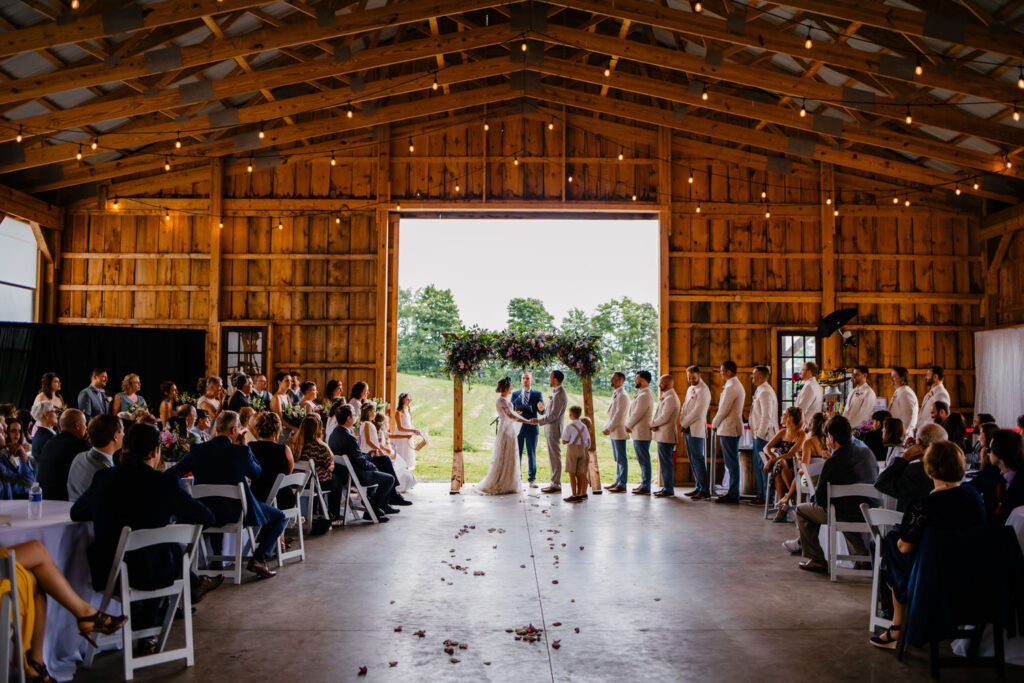 Guests are seated inside a wooden barn with open doors that frame the green landscape outside. The couple stands under a purple floral arch while the officiant conducts the ceremony.