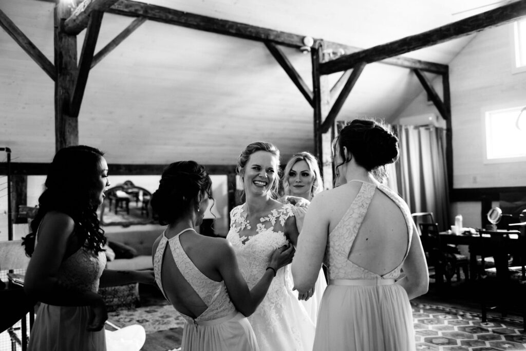 A bride laughs with her bridesmaids in a rustic room at The Treman Center, featuring exposed wooden beams.