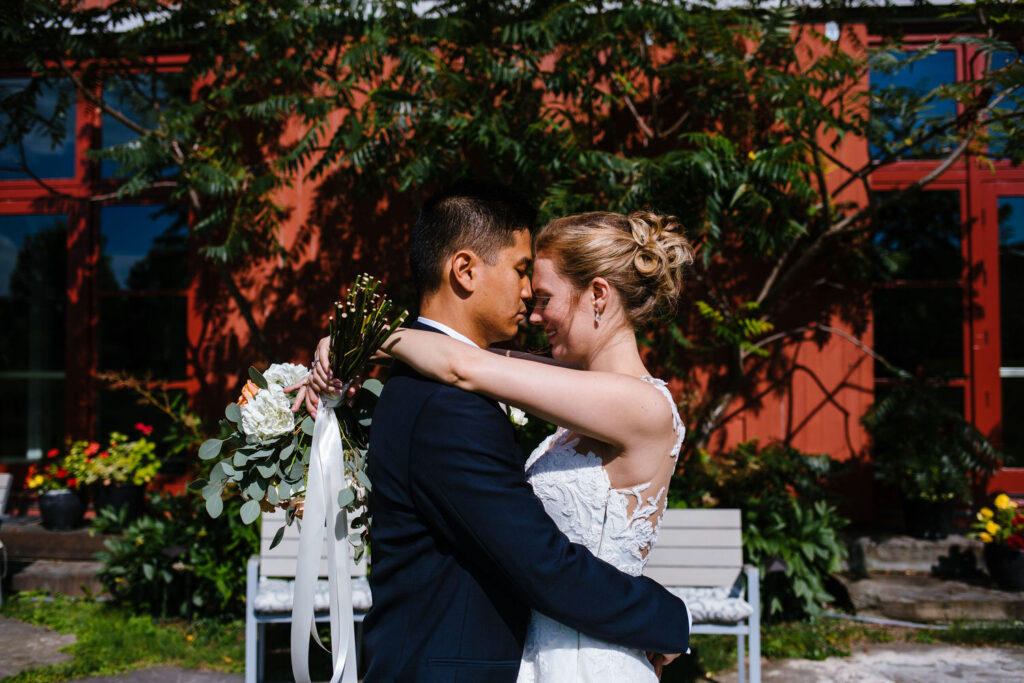 The bride and groom share a romantic moment in front of the red barn, with greenery and flowers.