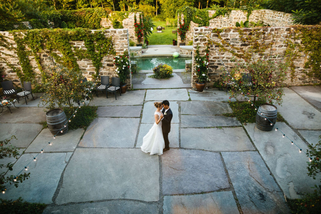 The bride and groom enjoy a romantic first dance in a stone courtyard, surrounded by ivy-covered walls and string lights at The Treman Center.