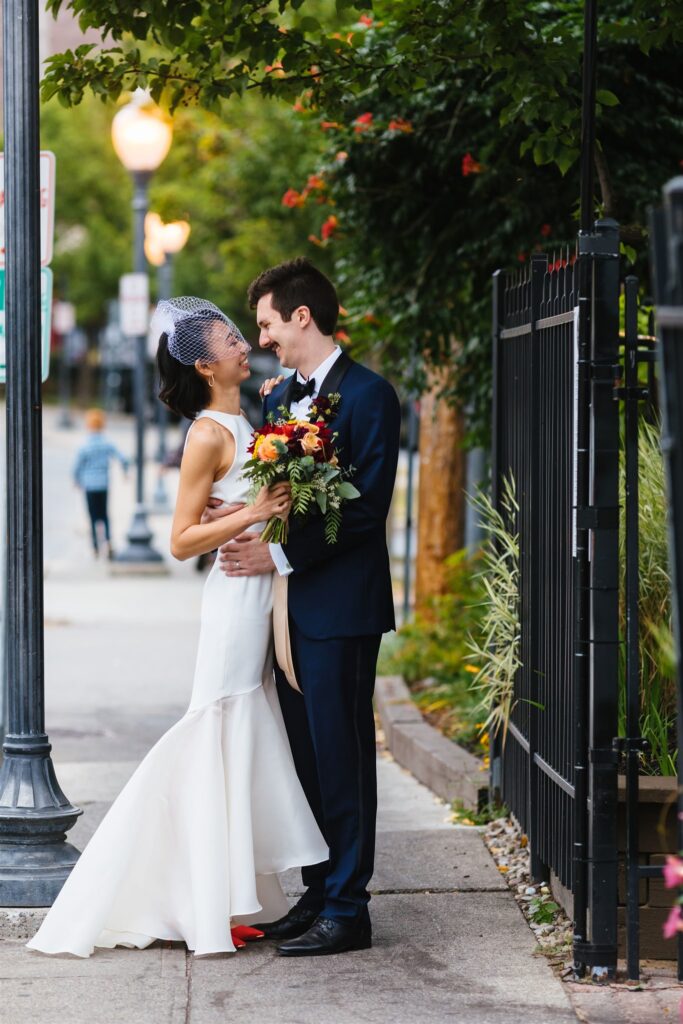Bride and groom embrace and smile at each other on the streets of Saratoga, NY. The bride holds a colorful bouquet of flowers.