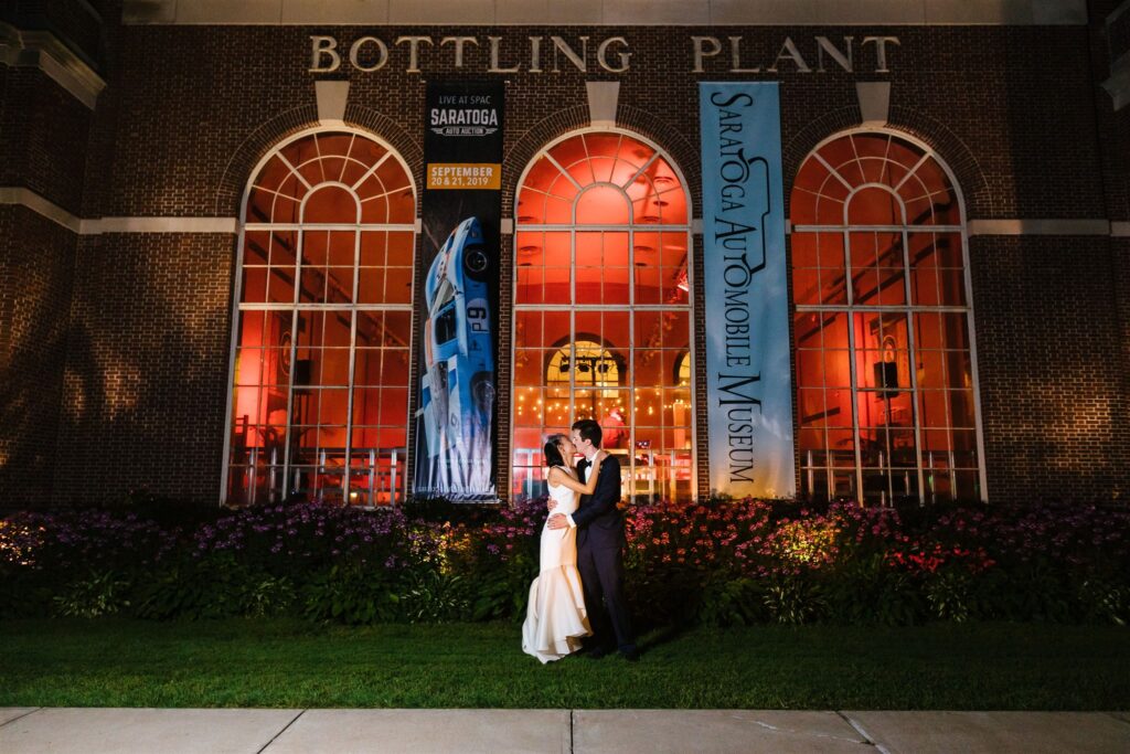 Bride and groom embrace and kiss in the evening in front of the Saratoga automobile museum where they had their wedding reception. 